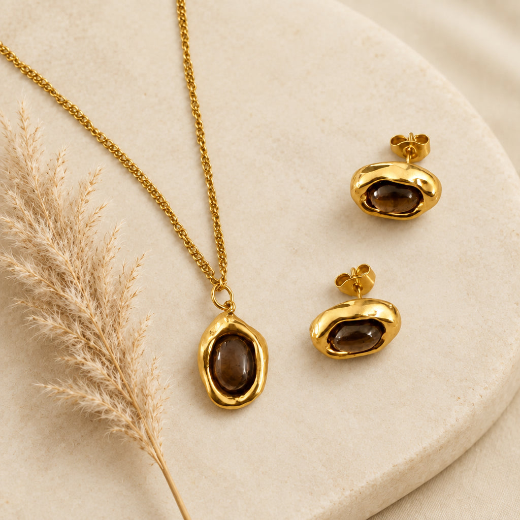 Close-up of a brown stone oval pendant in a wavy gold-tone setting with matching stud earrings on cream marble.