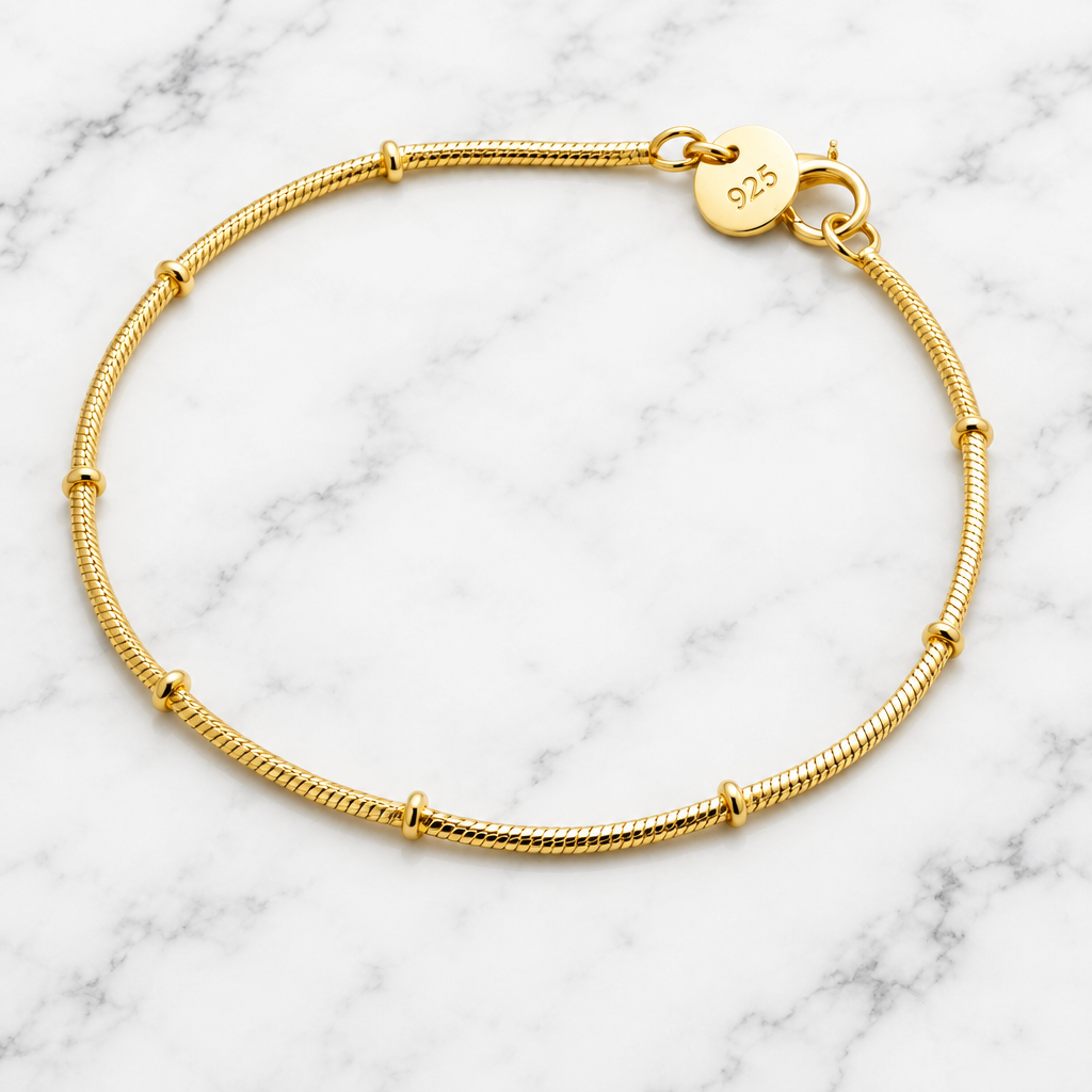 Thin gold-tone snake chain bracelet with small bamboo-style knots and a lobster claw clasp, flat lay on marble.