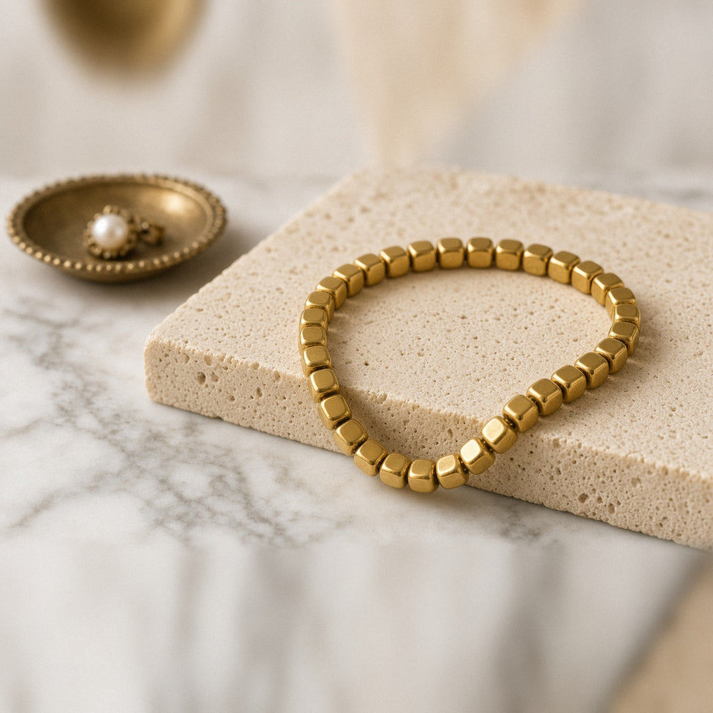 Gold-tone cube bead bracelet on a cream stone block beside a brass trinket dish