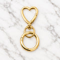 Gold-tone heart-shaped swivel key clip with a round spring ring below, on a marble surface.