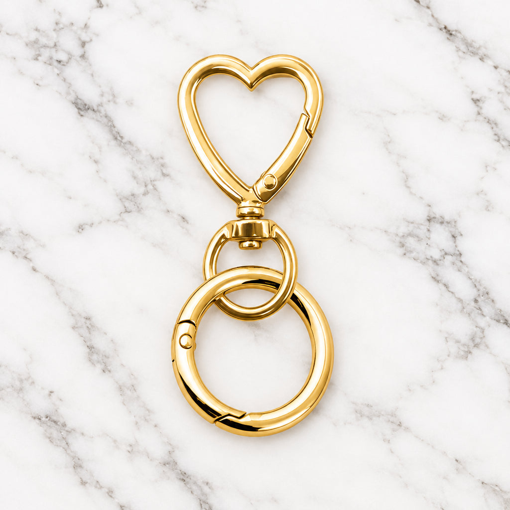 Gold-tone heart-shaped swivel key clip with a round spring ring below, on a marble surface.