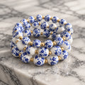 Stack of three stretch bracelets with white porcelain-style beads, dark blue floral pattern and small gold-tone spacer beads.