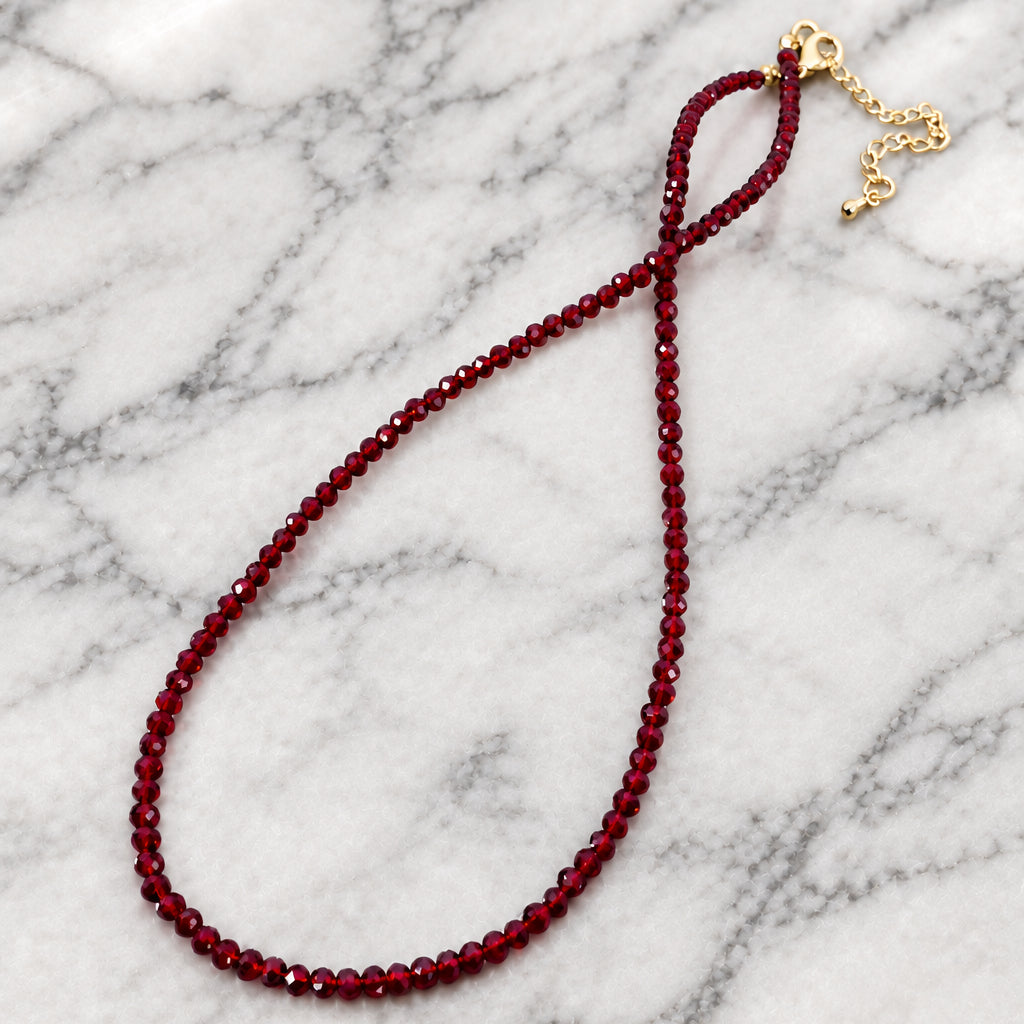 Single strand of small faceted deep red glass beads with a gold-tone lobster clasp and extender chain, laid in a loop on marble.