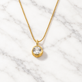 Gold-tone ball chain necklace with a round clear faceted stone in a polished gold bezel pendant, on marble.