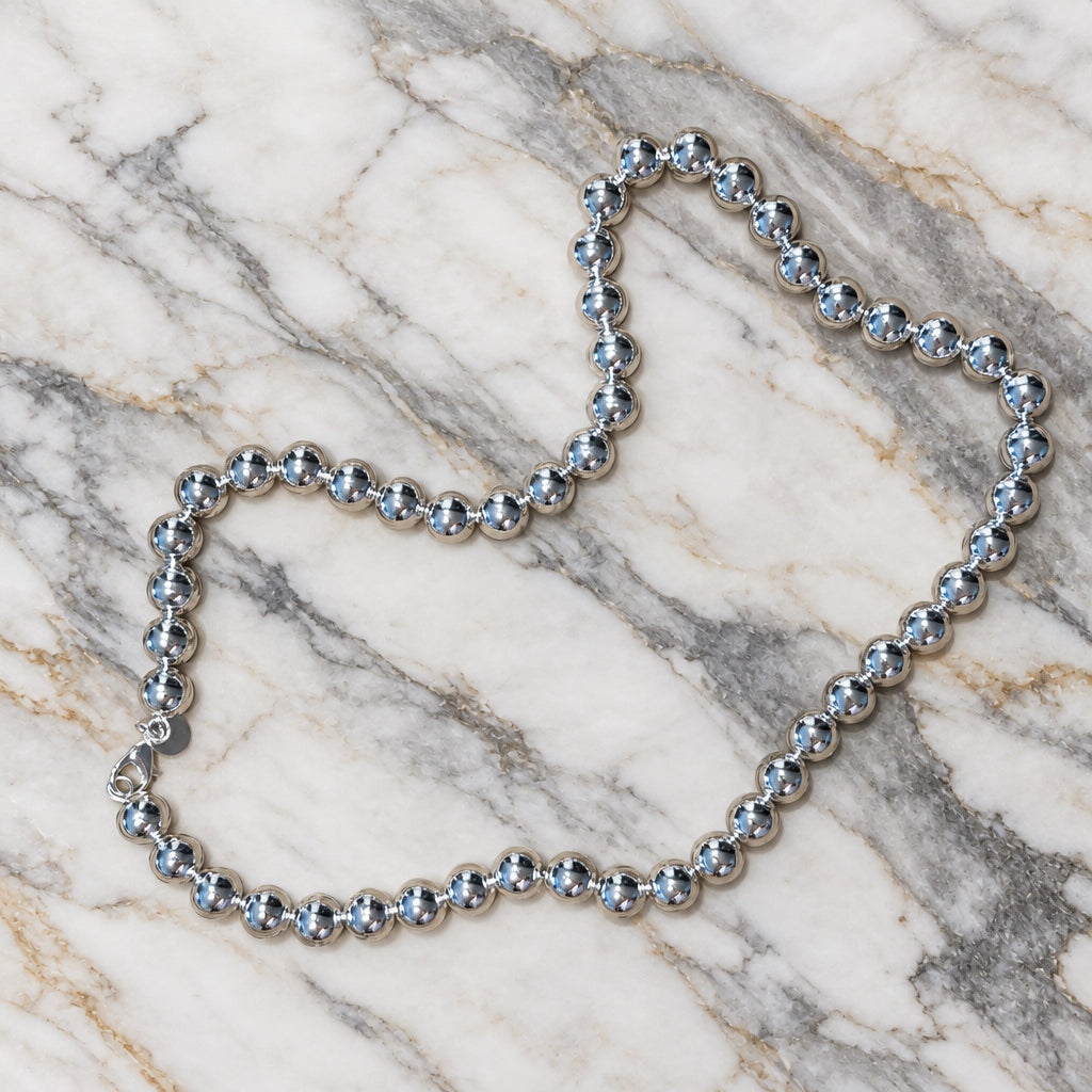 Sterling silver necklace of large polished round beads with a lobster clasp and small round tag, laid on marble.