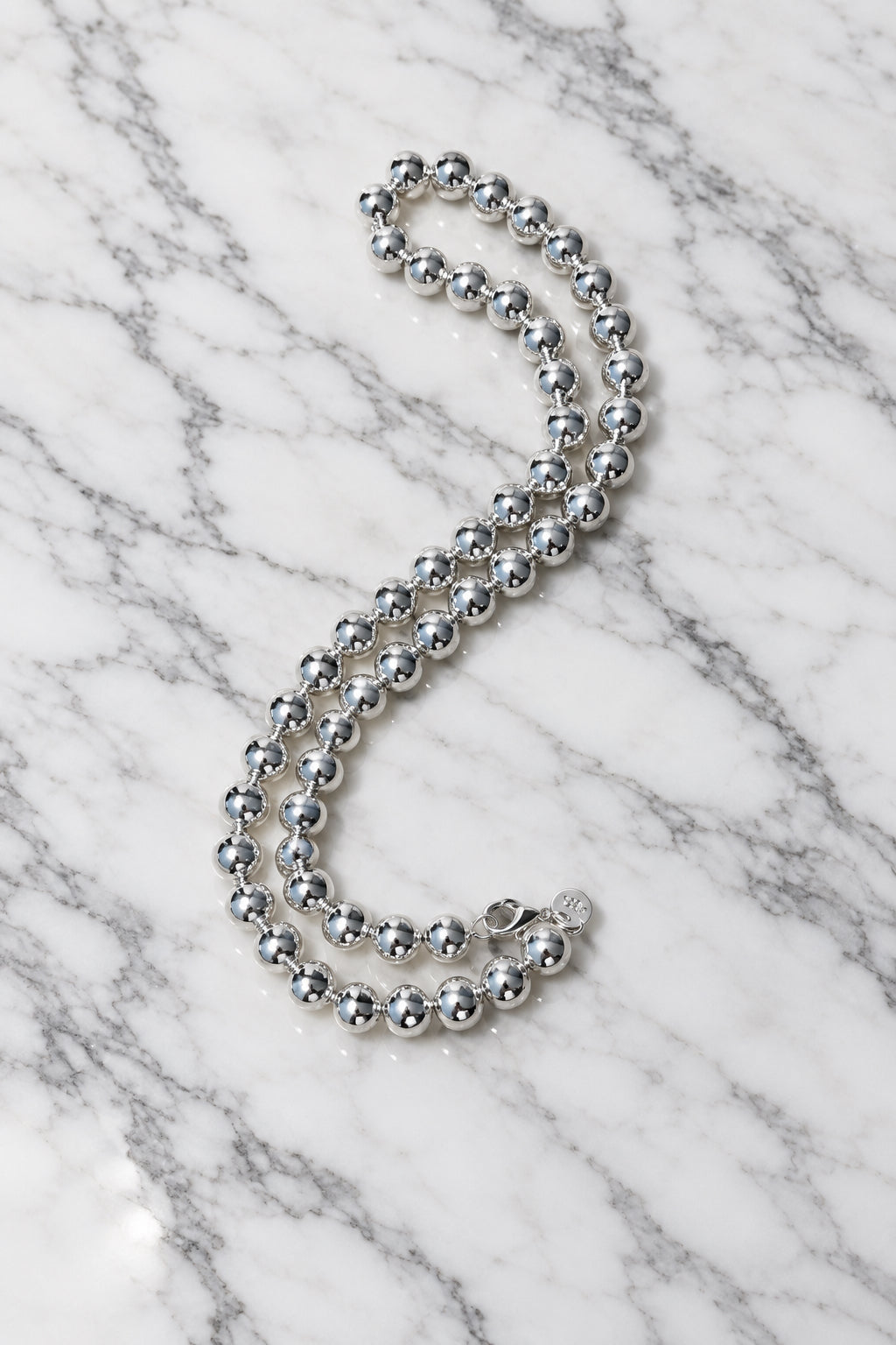Polished sterling silver ball bead necklace in an S-curve on white marble, showing the lobster clasp and round tag.