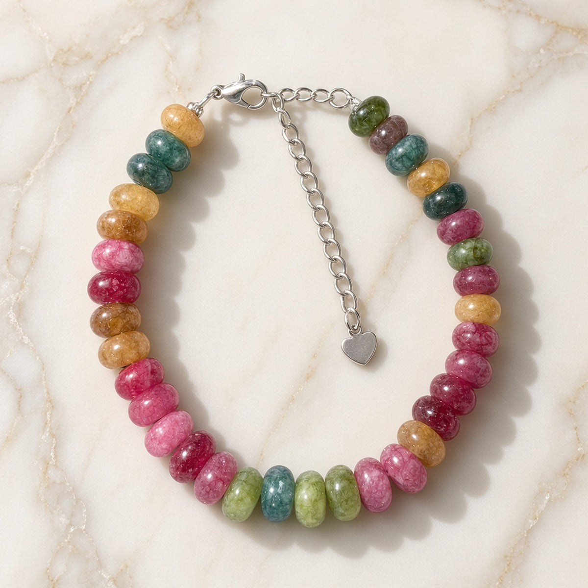 Pink, berry, green, and gold gemstone bracelet with silver heart charm on cream marble