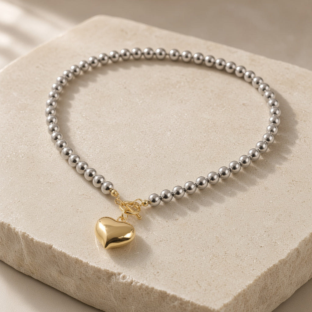 Silver bead necklace with gold heart pendant and toggle clasp on a cream stone block