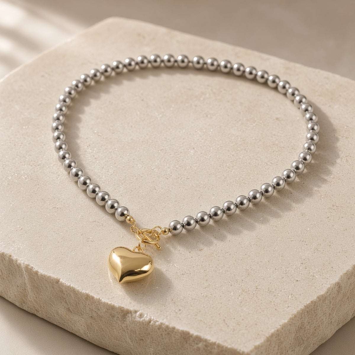 Silver bead necklace with gold heart pendant and toggle clasp on a cream stone block