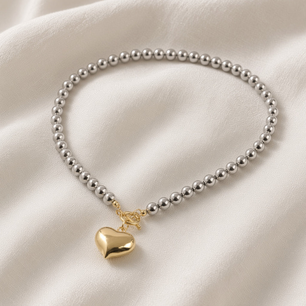 Silver bead necklace with gold heart pendant laid on soft draped ivory fabric