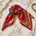 Red satin square scarf with gold chain, blue buckle strap, and medallion print, knotted on white marble.