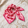 Pink and red heart print satin square scarf tied in a soft knot on gold-veined white marble.