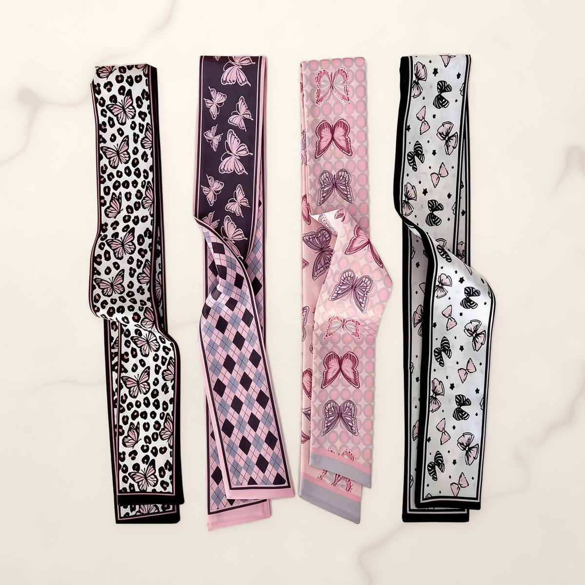 Four pink and black butterfly print twilly scarves with leopard, argyle, dot and bow accents on white marble.