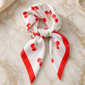 White satin square scarf with red cherry print and red border, tied in a soft knot on gold-veined marble.