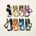 Eight folded twilly scarves with framed borders in black, chartreuse, taupe, lilac, navy, orange, blue and sage on white marble.