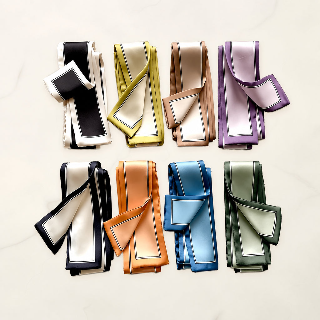 Eight folded twilly scarves with framed borders in black, chartreuse, taupe, lilac, navy, orange, blue and sage on white marble.