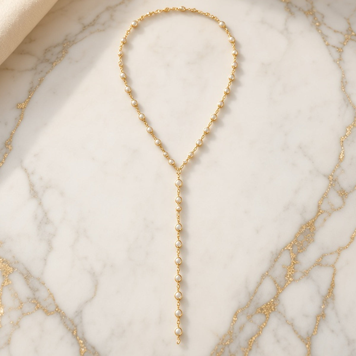 Long gold-tone Y-style lariat necklace with pearl-style beads set along the chain, laid flat on gold-veined marble.