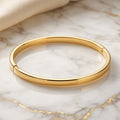 Glossy gold-plated sterling silver snap bangle with a hinged closure, resting on gold-veined marble.