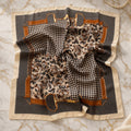 Black and cream houndstooth satin square scarf with paisley center, caramel buckle-strap print, and cream border, laid flat on gold-veined marble.