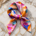 Satin square scarf with abstract pink, orange, blue and lilac petal print, tied in a soft knot on gold-veined marble.