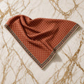 Rust red twill triangle scarf with tiny cream diamond print and black floral border, casually laid on gold-veined marble.