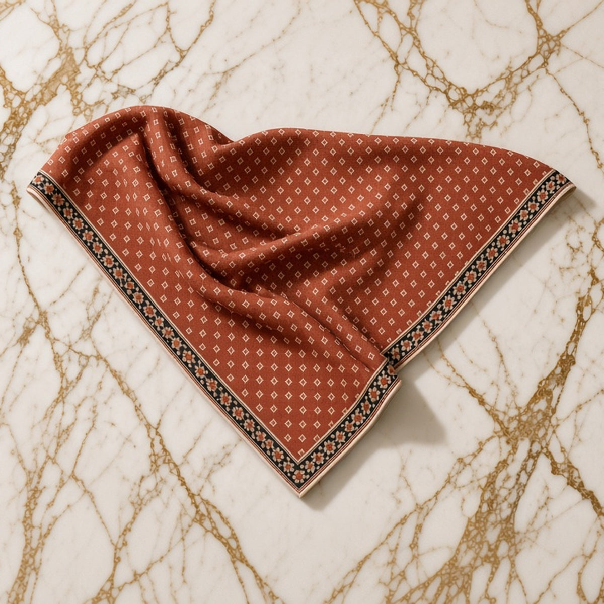 Rust red twill triangle scarf with tiny cream diamond print and black floral border, casually laid on gold-veined marble.
