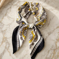 Cream satin square scarf with gray and mustard medallion print and black border, tied in a soft knot on gold-veined marble.