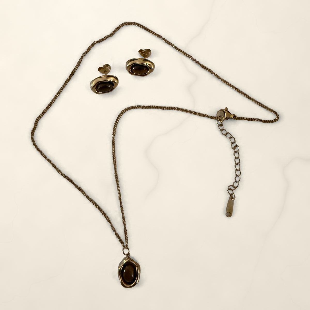 Gold chain necklace with a brown stone oval pendant and matching stud earrings on cream marble.