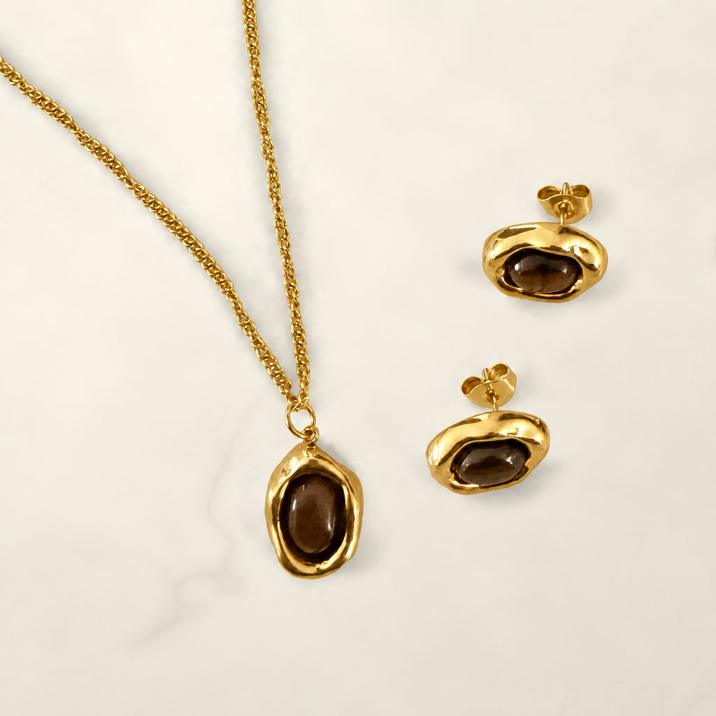 Close-up of a brown stone oval pendant in a wavy gold-tone setting with matching stud earrings on cream marble.