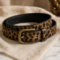 Slim leopard print faux leather belt with antique brass buckle coiled on cream linen.