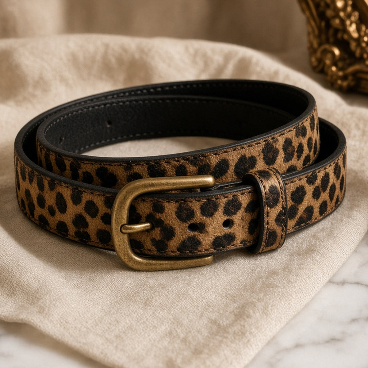 Slim leopard print faux leather belt with antique brass buckle coiled on cream linen.